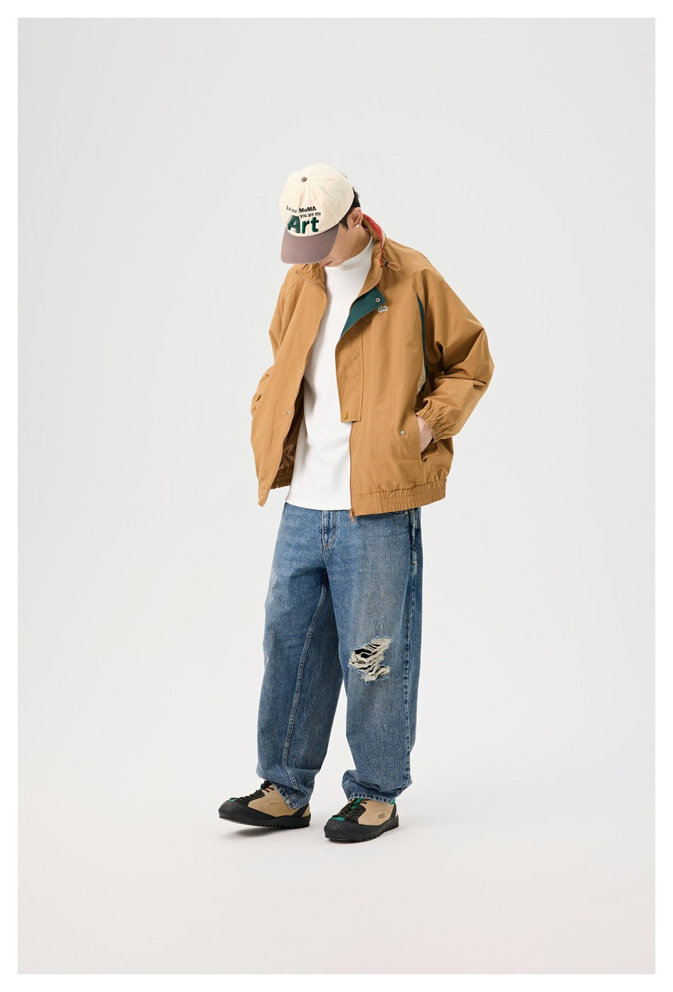 Model wearing the tan color-block jacket with hands in pockets, head tilted down, with a cap and wide-leg jeans