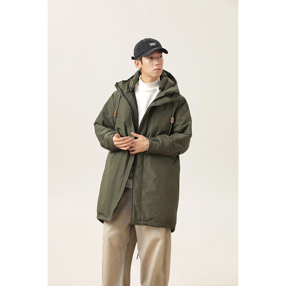 Model wearing a long dark olive hooded parka with orange cord toggles, shown from the front.