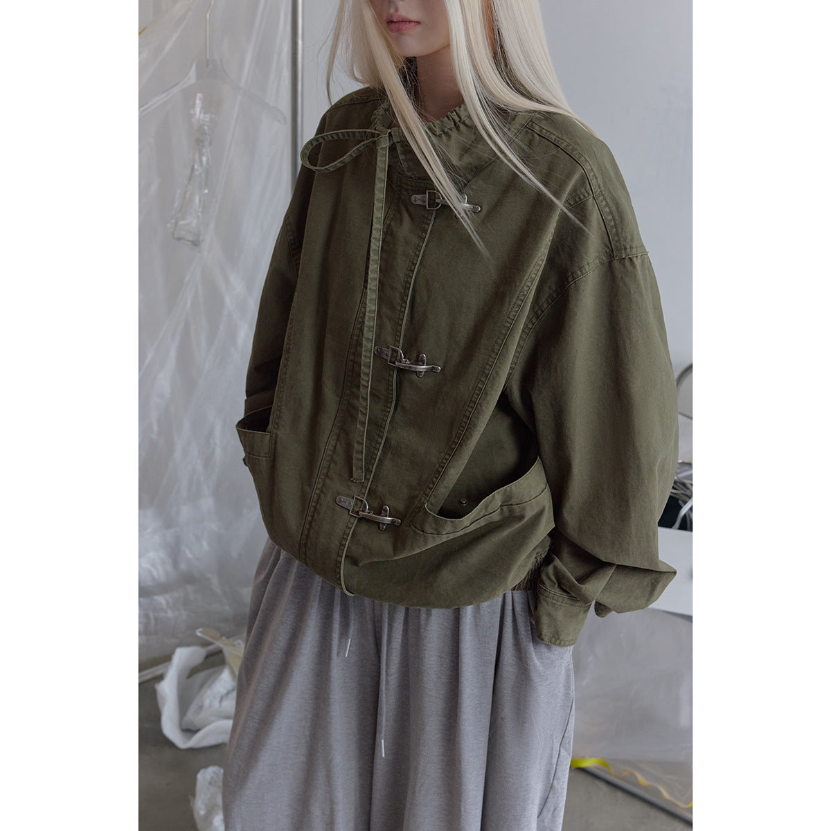 Close view of a model wearing an olive jacket with silver hook closures, a drawstring collar, and large front pockets.