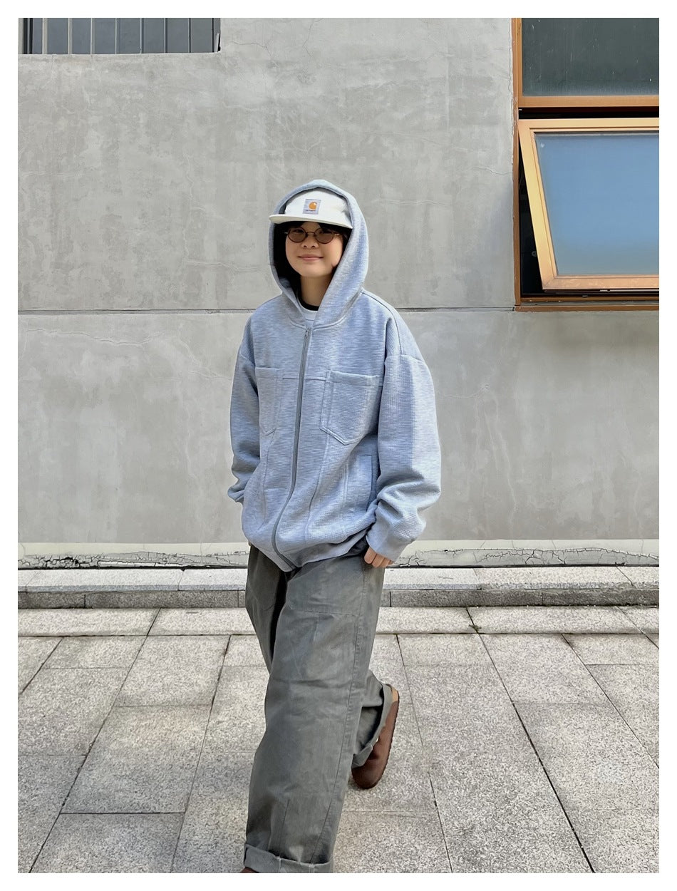 Person wearing a light gray hooded zip jacket over loose gray pants, with the hood up.