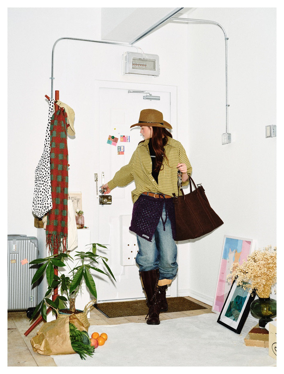 Model in a yellow checked shirt and blue jeans, carrying a large brown tote; three-quarter view.