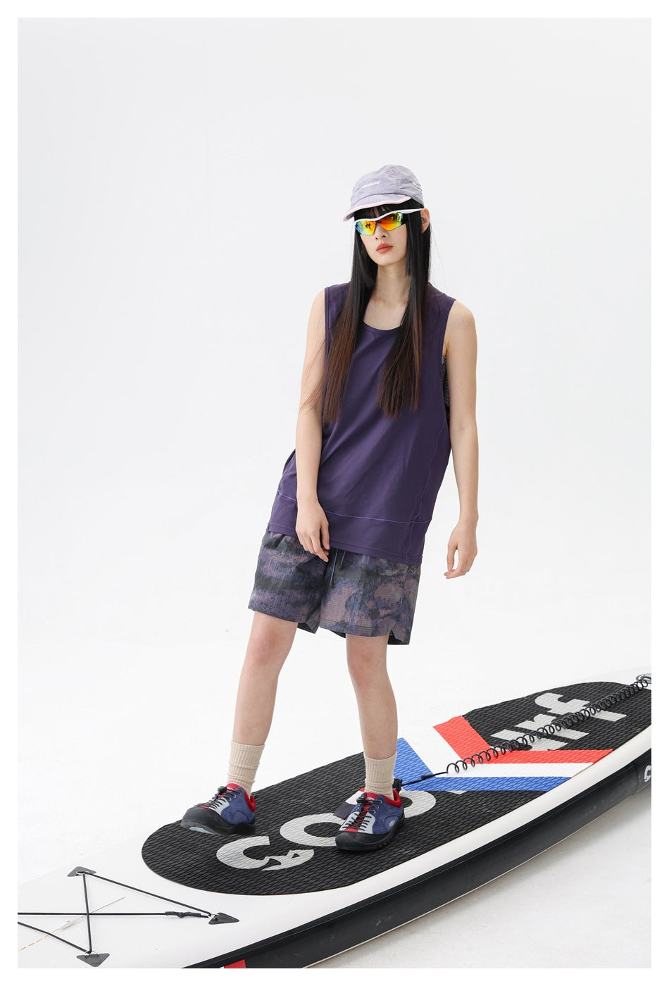 Model wearing a dark purple sleeveless top and muted purple-gray patterned shorts, standing on a paddleboard.