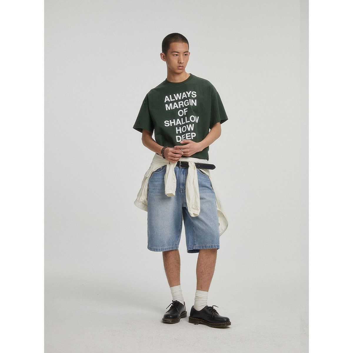 Model wearing a dark green graphic T-shirt, light blue denim shorts, and a cream jacket tied at the waist.