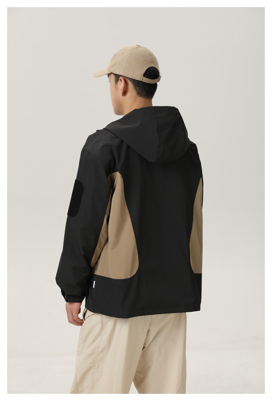 Rear view of a model wearing a black hooded jacket with tan side panels and beige trousers.