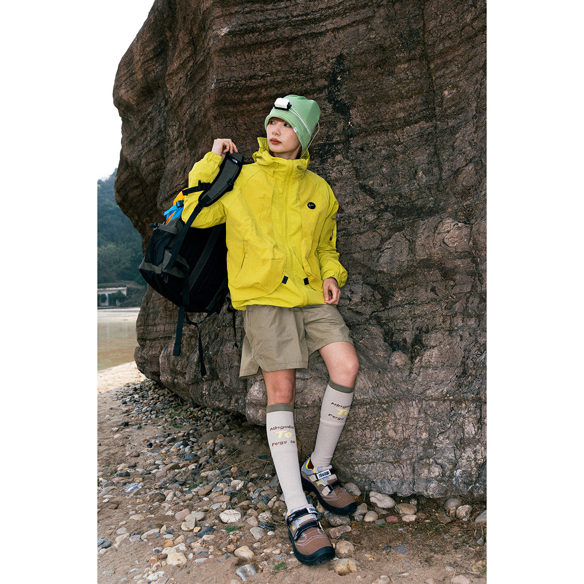 A person models a bright yellow hooded outdoor jacket with beige shorts beside a rock face.