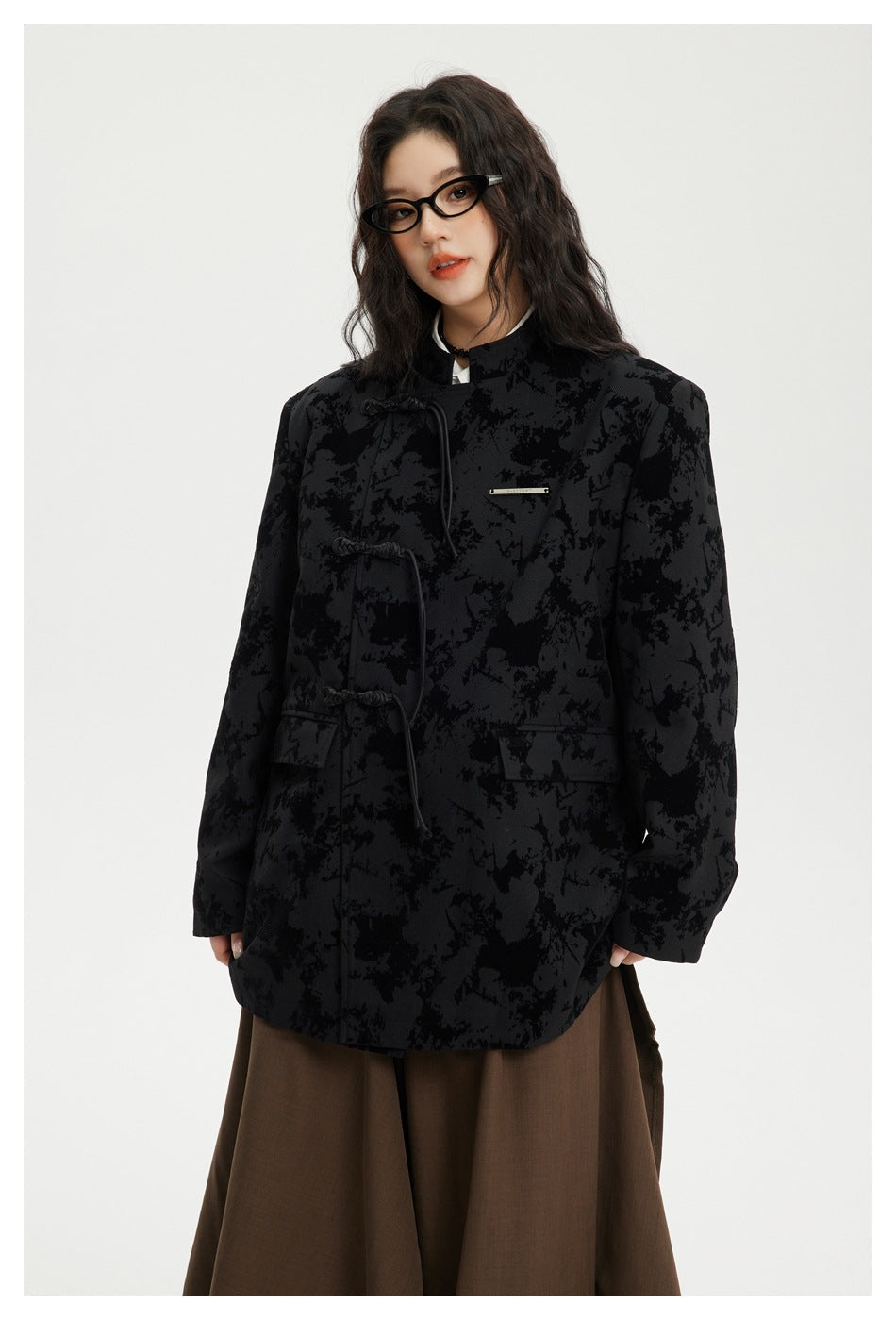Front view of a model wearing a black abstract-patterned jacket with cord toggles and flap pockets over brown trousers.