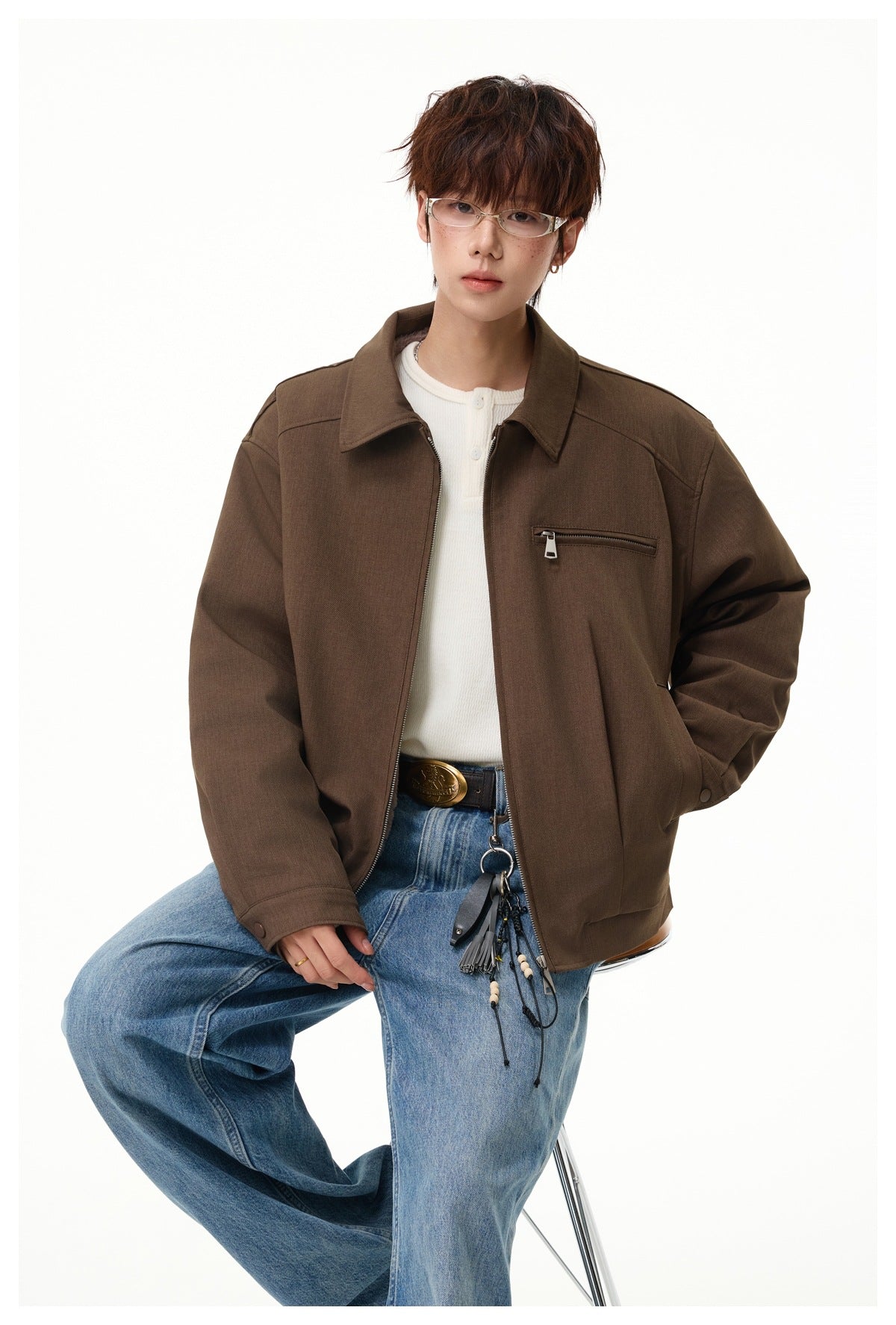 Model seated in a brown zip-front jacket with a pointed collar and chest zip pocket, paired with a white top and jeans.