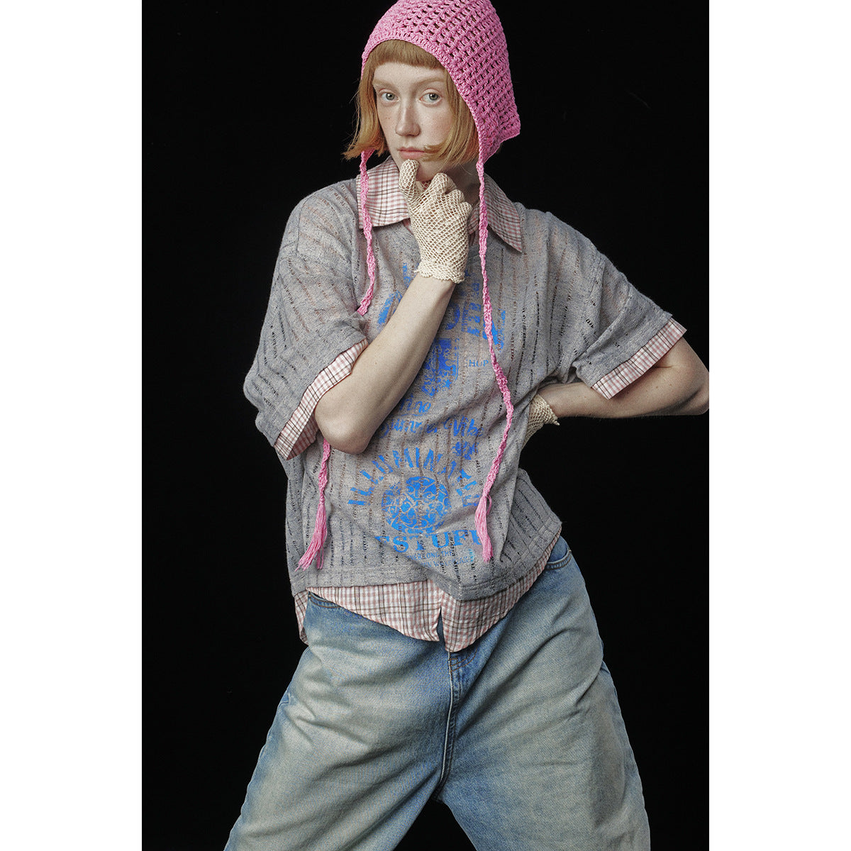 Model wearing a gray blue-graphic tee over a checked shirt, loose jeans, a pink crochet bonnet, and cream crochet gloves.