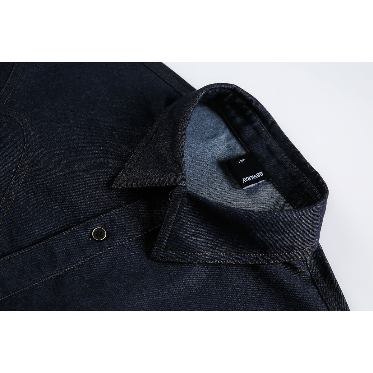 Close-up of a dark indigo denim garment showing a pointed collar, button, contrast stitching, and black neck label.