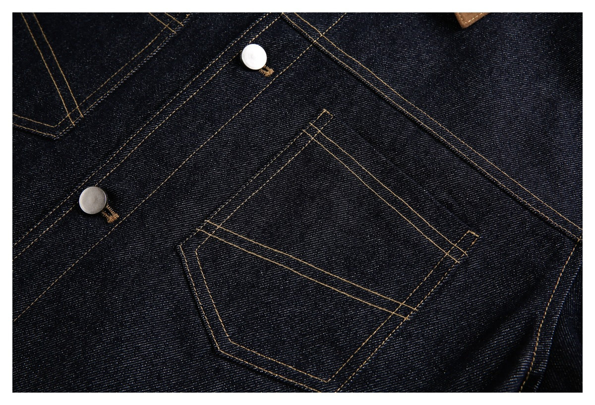 Dark indigo denim close-up with a stitched pocket, tan seams, metal buttons, and a brown collar edge.