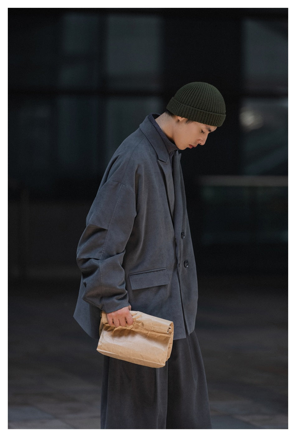 Side view of a model wearing an oversized slate-gray coat, dark trousers, and an olive ribbed beanie.