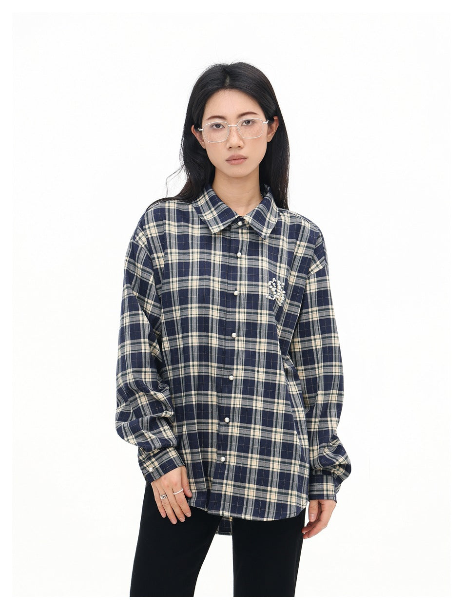 Model wearing a buttoned navy and cream plaid shirt with pearl-like chest embellishment and black trousers.