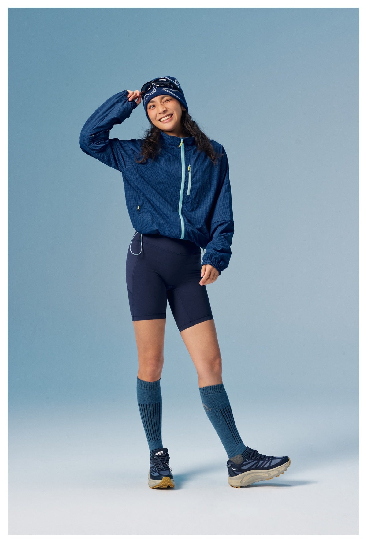 Model wearing a navy zip-front jacket and dark cycling shorts, with blue knee socks and trail shoes.
