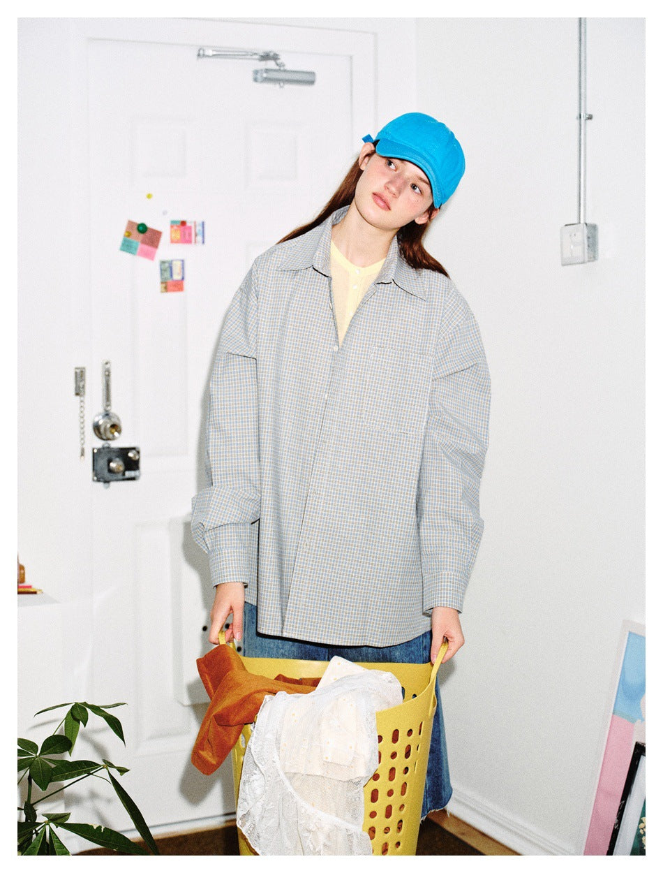 Model wearing an oversized gray-blue plaid button-up shirt and bright blue cap, holding a laundry basket.