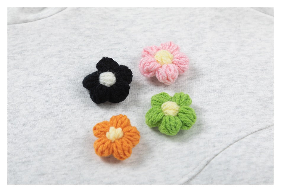 Four raised yarn flowers in black, pink, green, and orange decorate light heather-gray fabric.