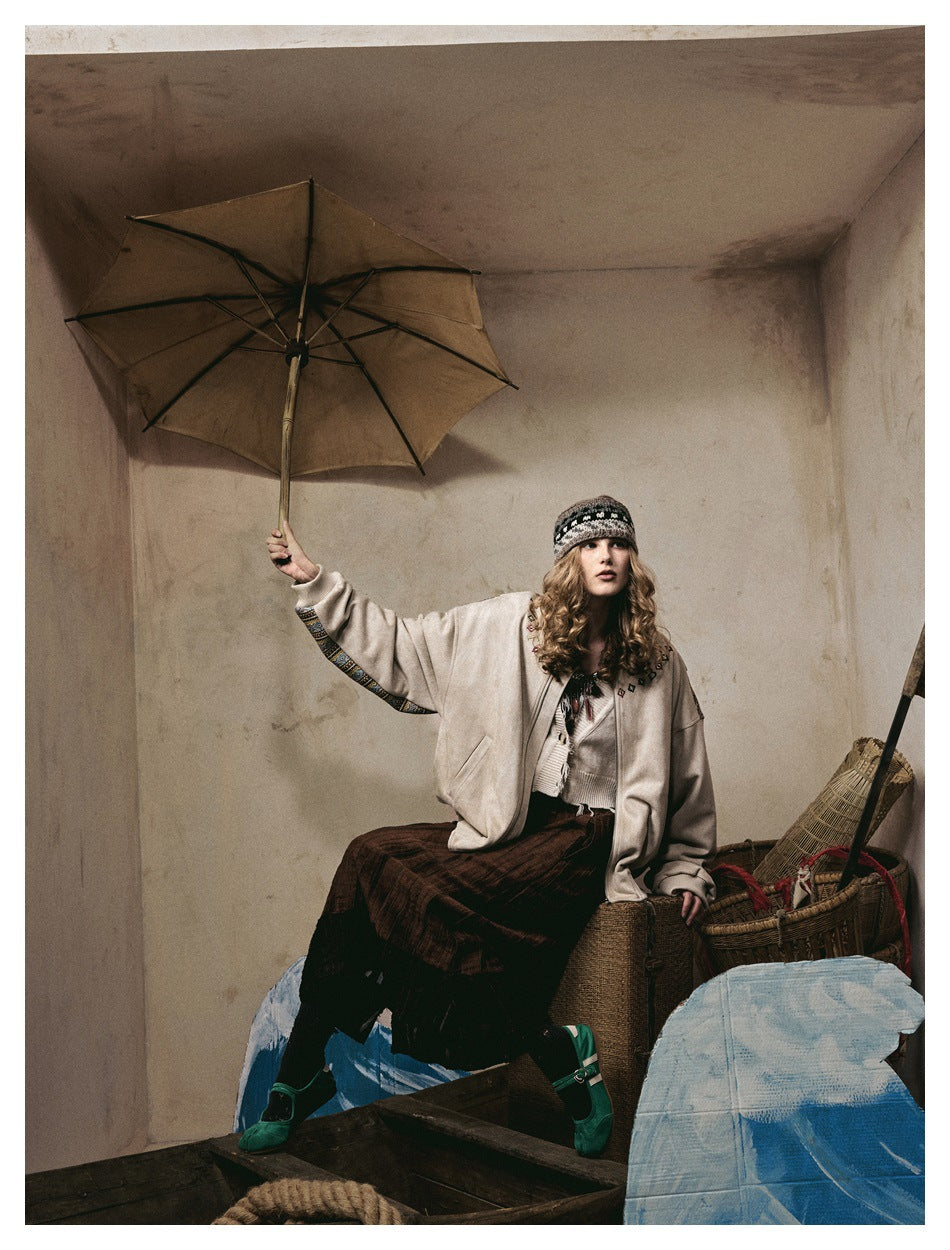 Model seated in a cream jacket with patterned sleeves and embroidered chest details, paired with a knit top and dark skirt.