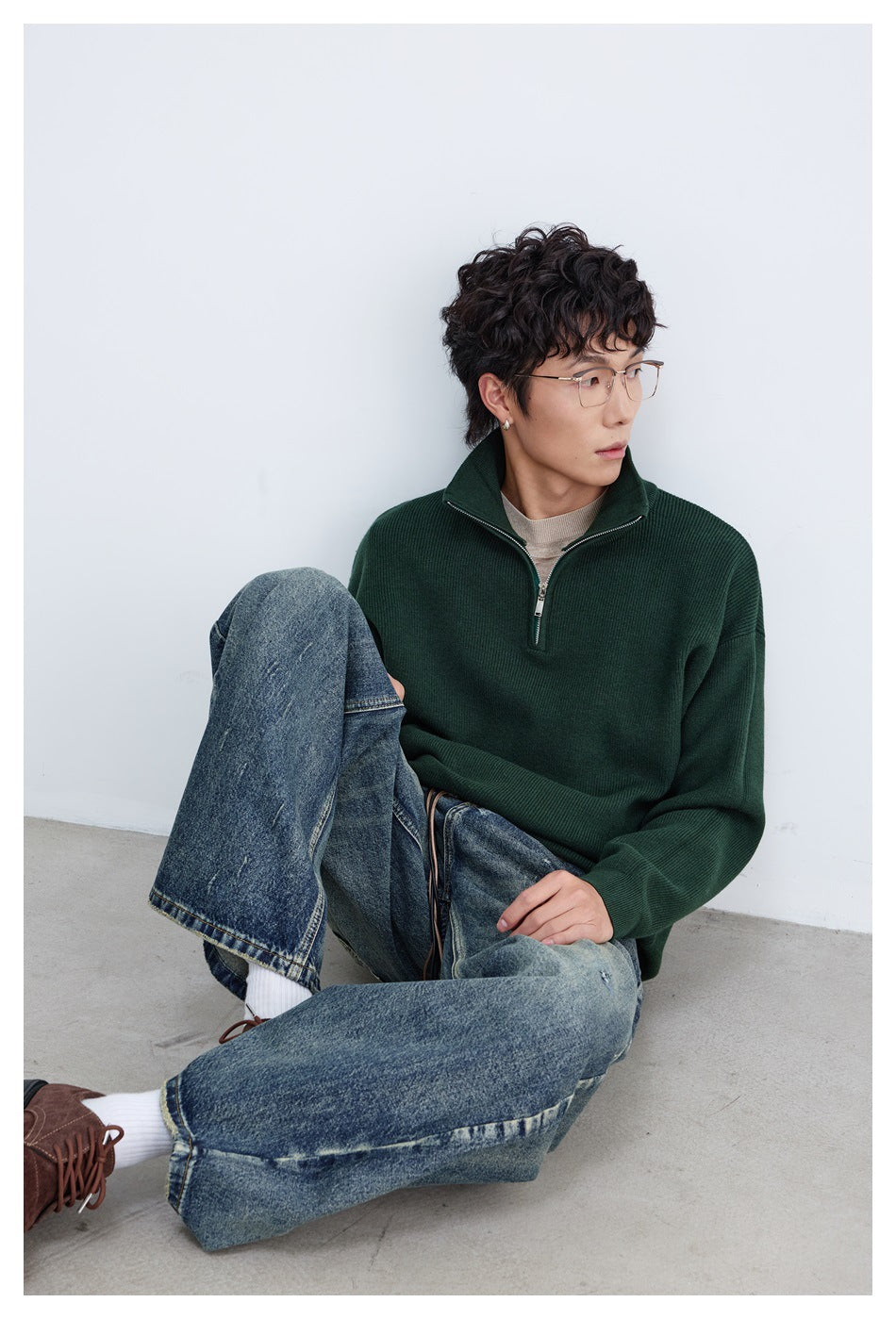 Seated model wearing a dark green quarter-zip pullover and relaxed blue jeans.