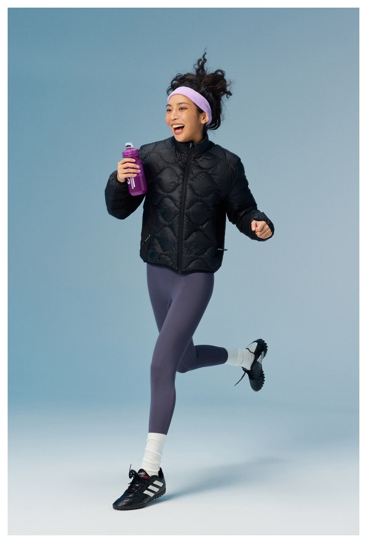 Woman jogging in the black quilted stand-collar jacket with leggings and a purple headband, holding a water bottle