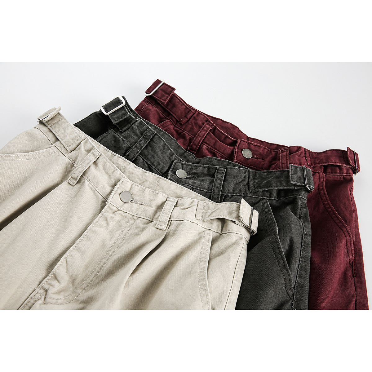 Close-up of beige, charcoal, and burgundy cargo pants showing adjustable waist tabs and front pockets.