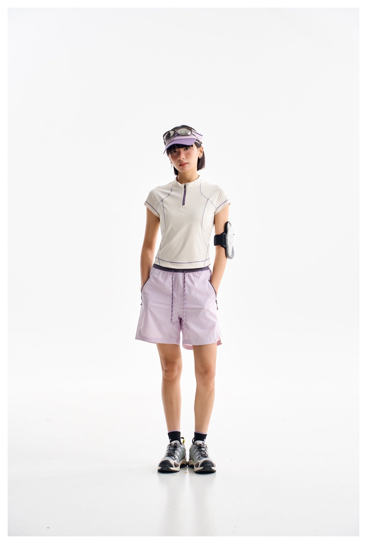 Model wearing the off-white half-zip short-sleeve top with purple stitching, styled with lavender shorts and a visor