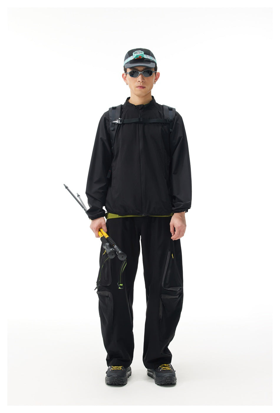 Model wearing a black hiking jacket and cargo pants with a cap, sunglasses, backpack straps and trekking poles.