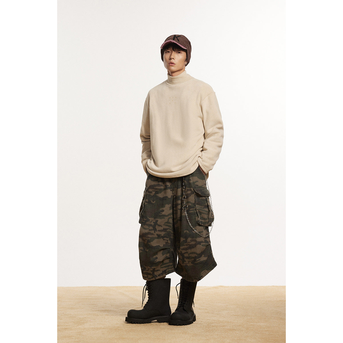 Full-length model wearing a cream high-neck pullover, camouflage cargo shorts, black boots, and a brown cap.