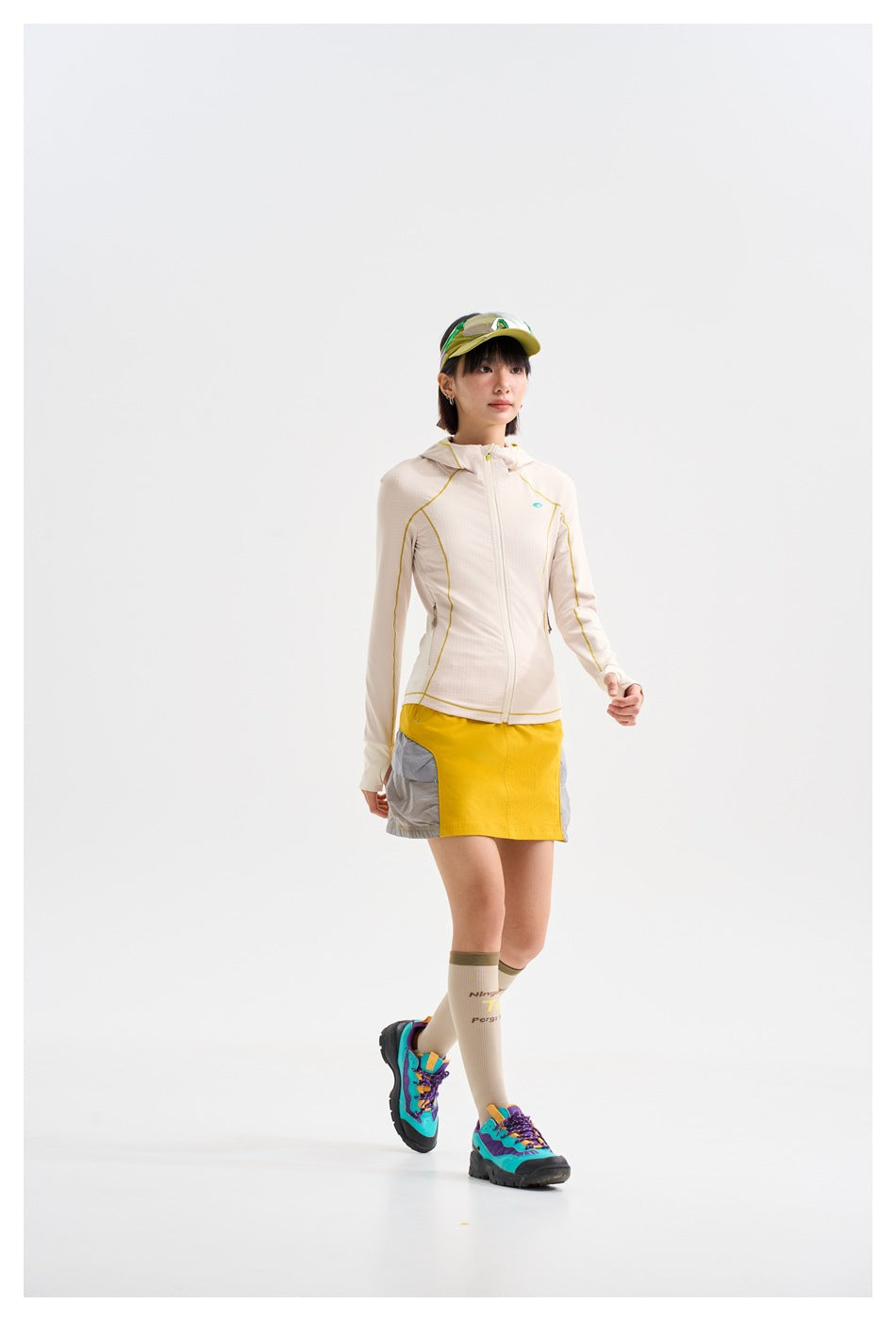 Model walking in a cream hooded jacket with yellow piping and a yellow-and-gray skirt, with a cap, knee socks and colorful