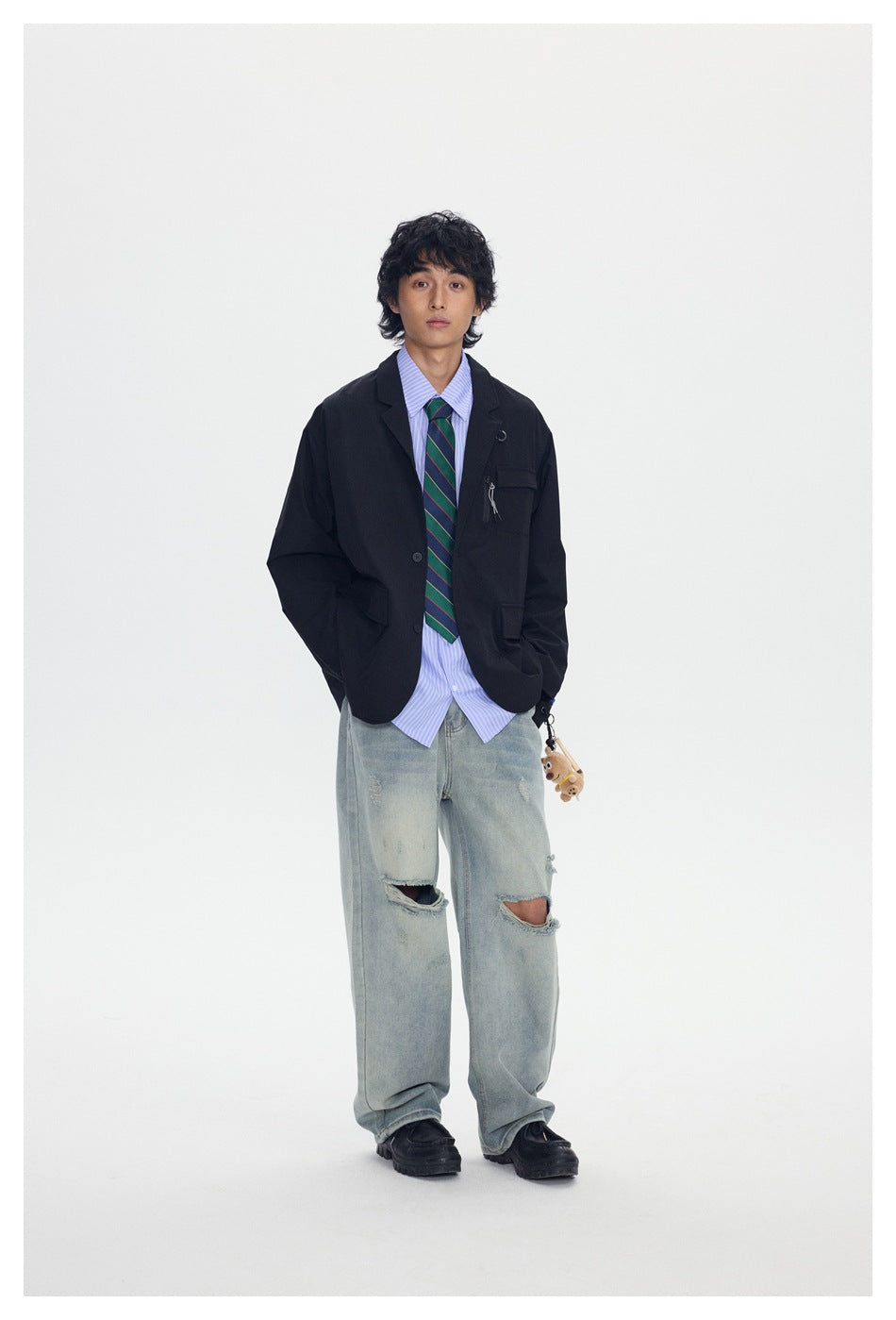 Model with hands in pockets wearing a black relaxed jacket, striped shirt, green tie and ripped jeans