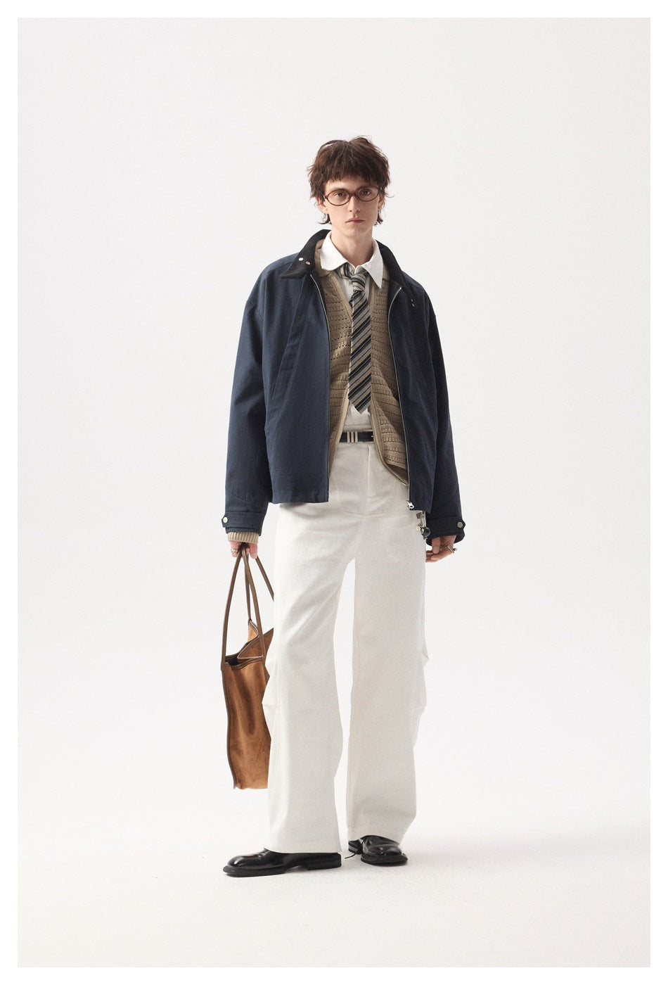 Full-length model wearing a navy jacket over a knit vest, shirt and tie with white trousers.