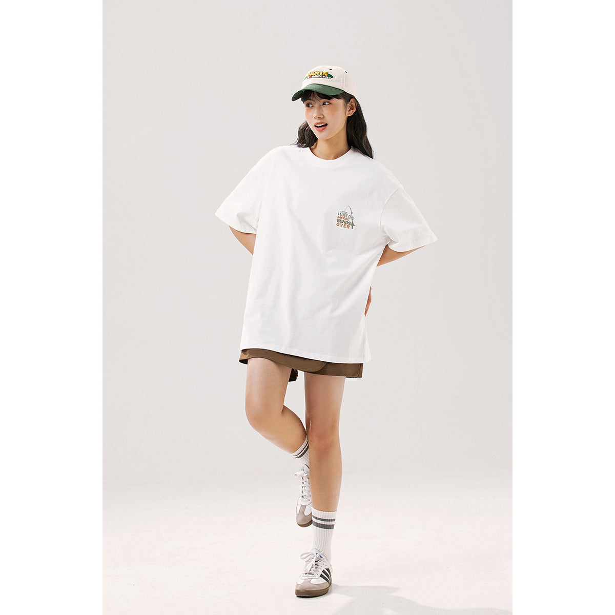 Model wearing an oversized white T-shirt with a small multicolor chest graphic, brown shorts, and a cap.