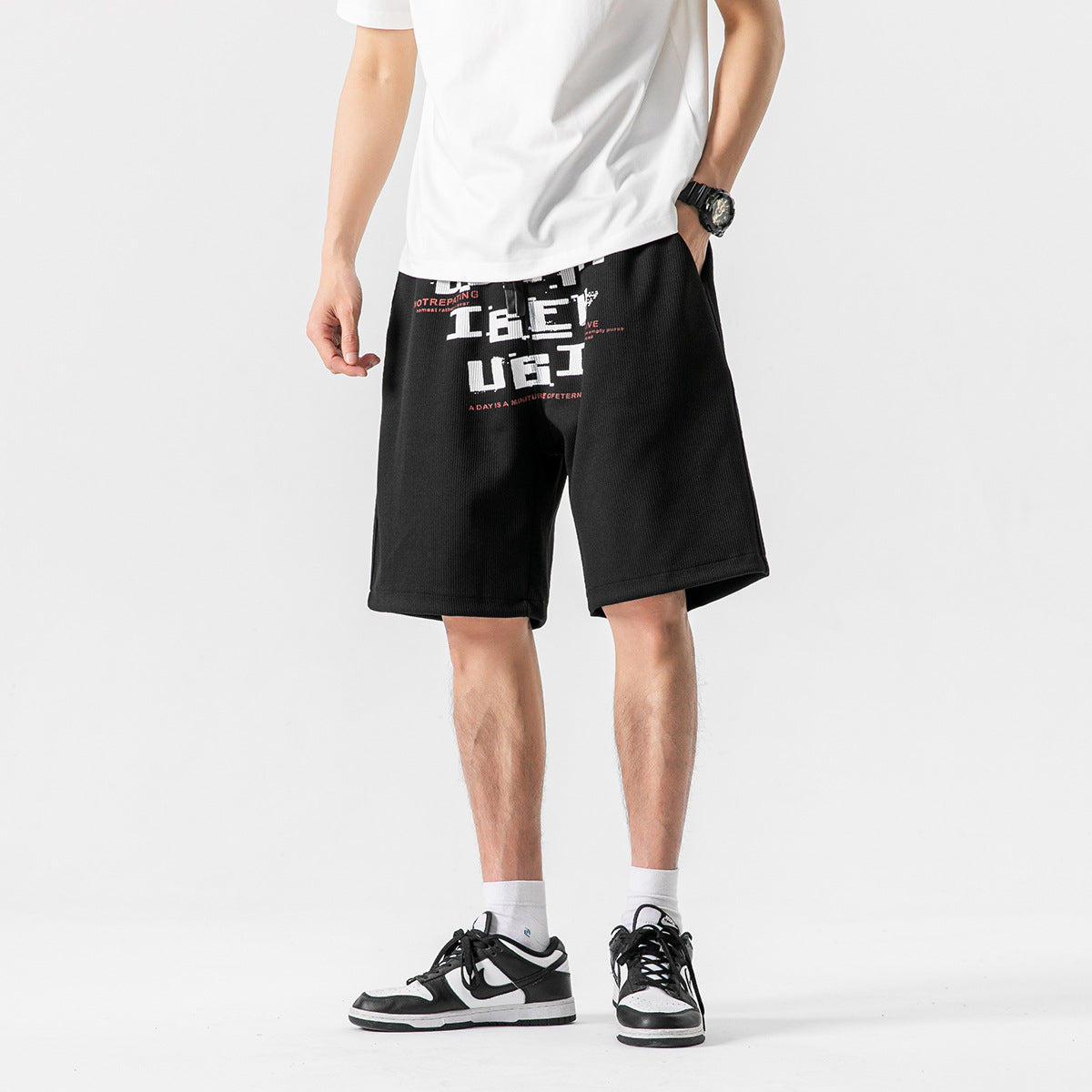 Front view of a model wearing black knee-length shorts with a bold white and red text graphic.