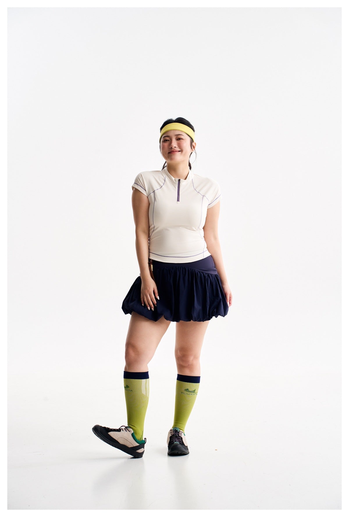 Full-length model wearing an ivory zip-neck top, navy bubble skirt, lime knee-high socks, and a yellow headband.