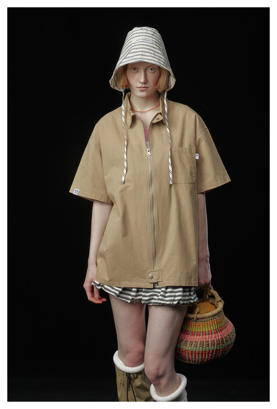 Front view of a model in an oversized tan short-sleeve zip jacket, striped hat and shorts.