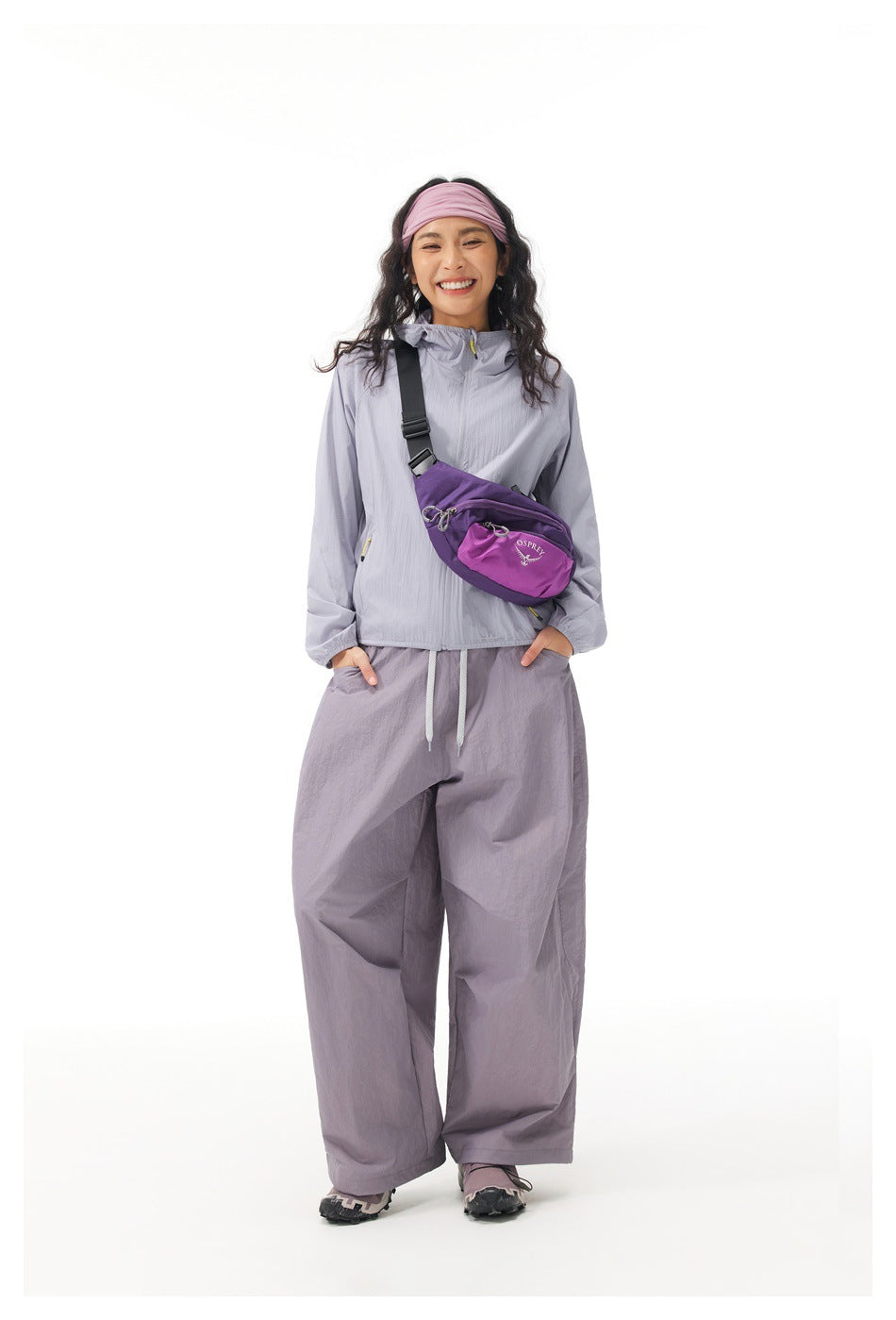 Full-length model wearing a light gray-lavender jacket and loose trousers, with a purple crossbody waist bag.