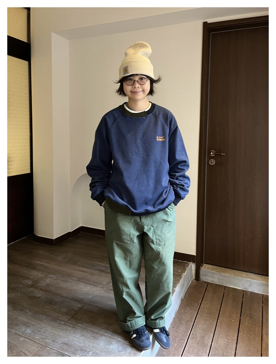 Person wearing a navy crewneck sweatshirt with contrast trim, loose green trousers, and a cream beanie.