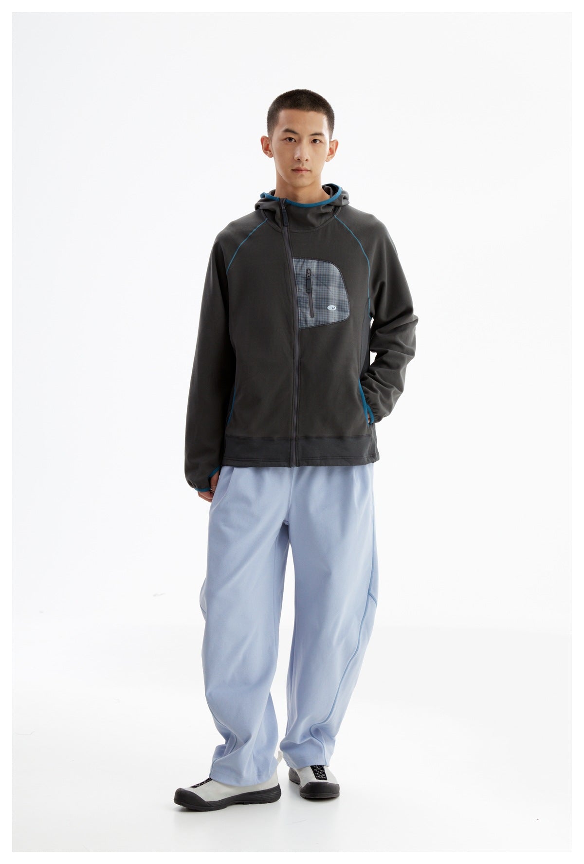 Full-length model wearing a charcoal hooded zip-up jacket with blue piping and a plaid chest pocket, plus loose light-blue