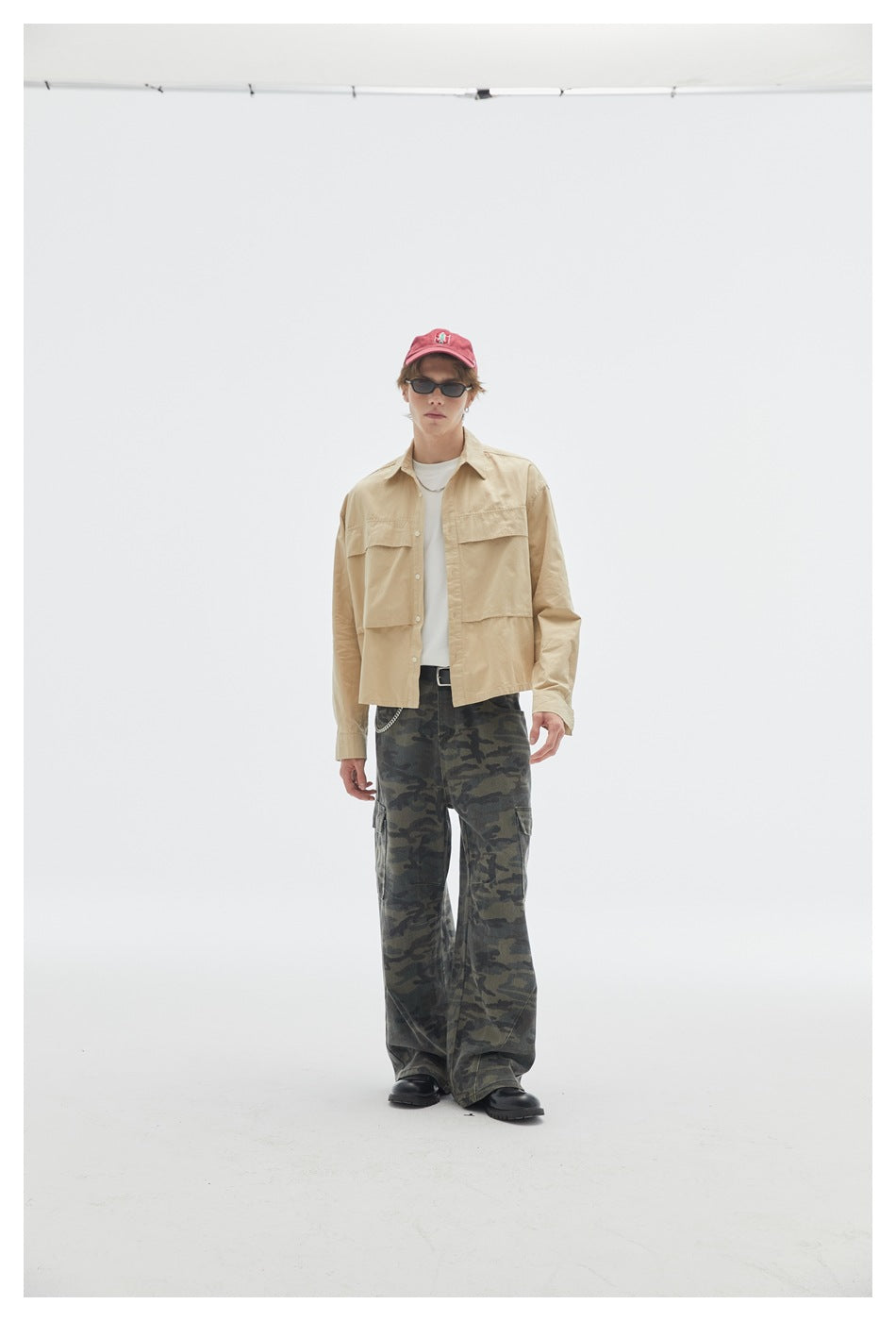 Model in a beige button-up overshirt and loose dark camouflage cargo pants, standing at a slight angle.