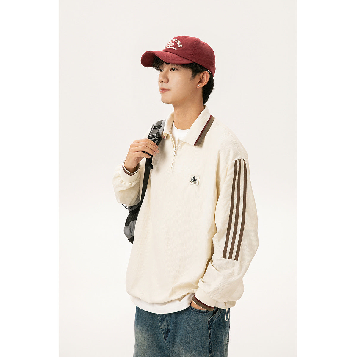 Model wearing a cream quarter-zip collared top with brown sleeve stripes and trim, paired with a red cap and backpack.