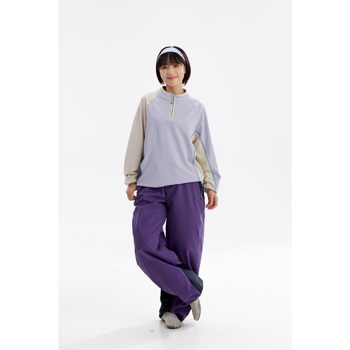 Model wearing a light lavender quarter-zip top with beige sleeves and loose purple pants, posing with crossed legs.