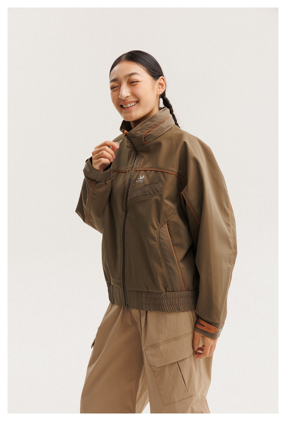 Model wearing a brown zip-front jacket with orange piping, a high collar, chest pocket and elastic hem.