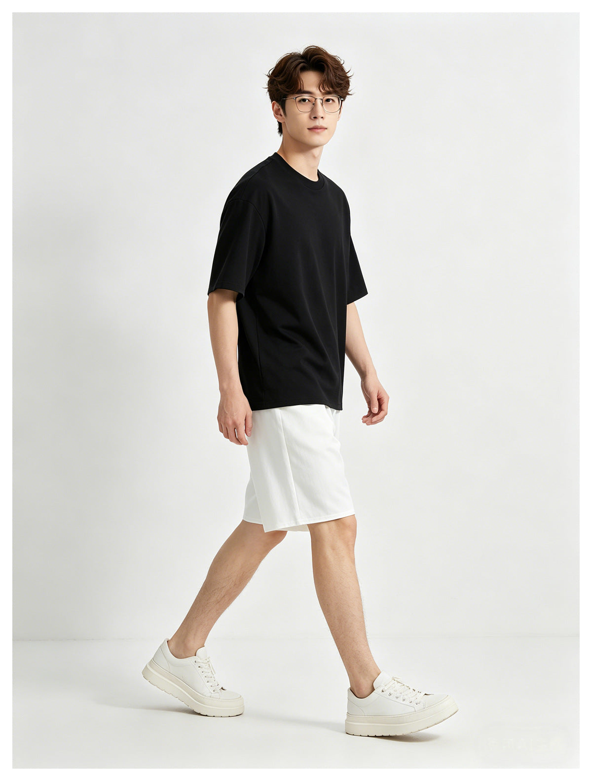 Model wearing a plain black oversized T-shirt, white shorts, and white sneakers, shown walking against a light backdrop.