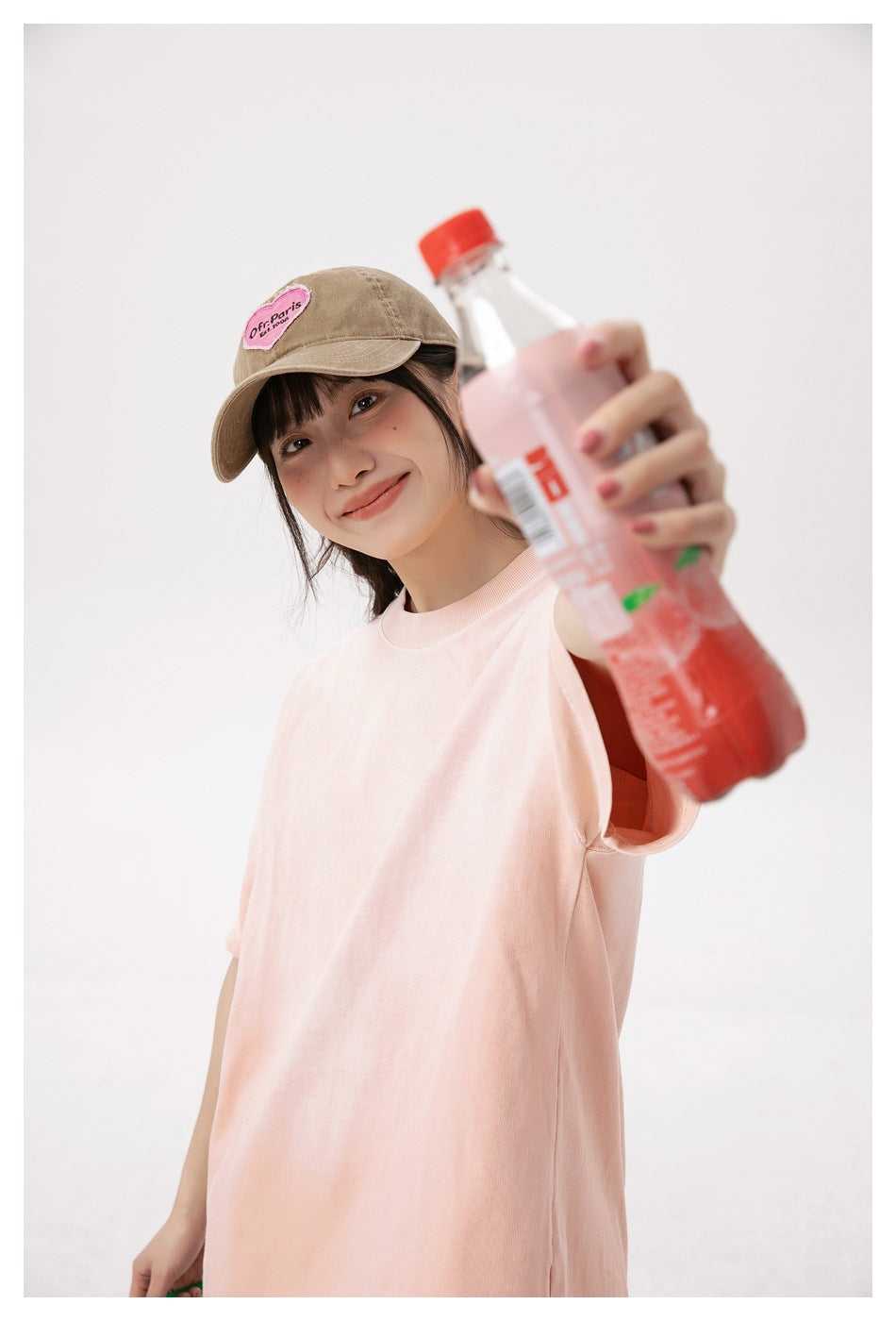 Close-up of a model in a pink T-shirt and brown cap holding a bottle toward the camera