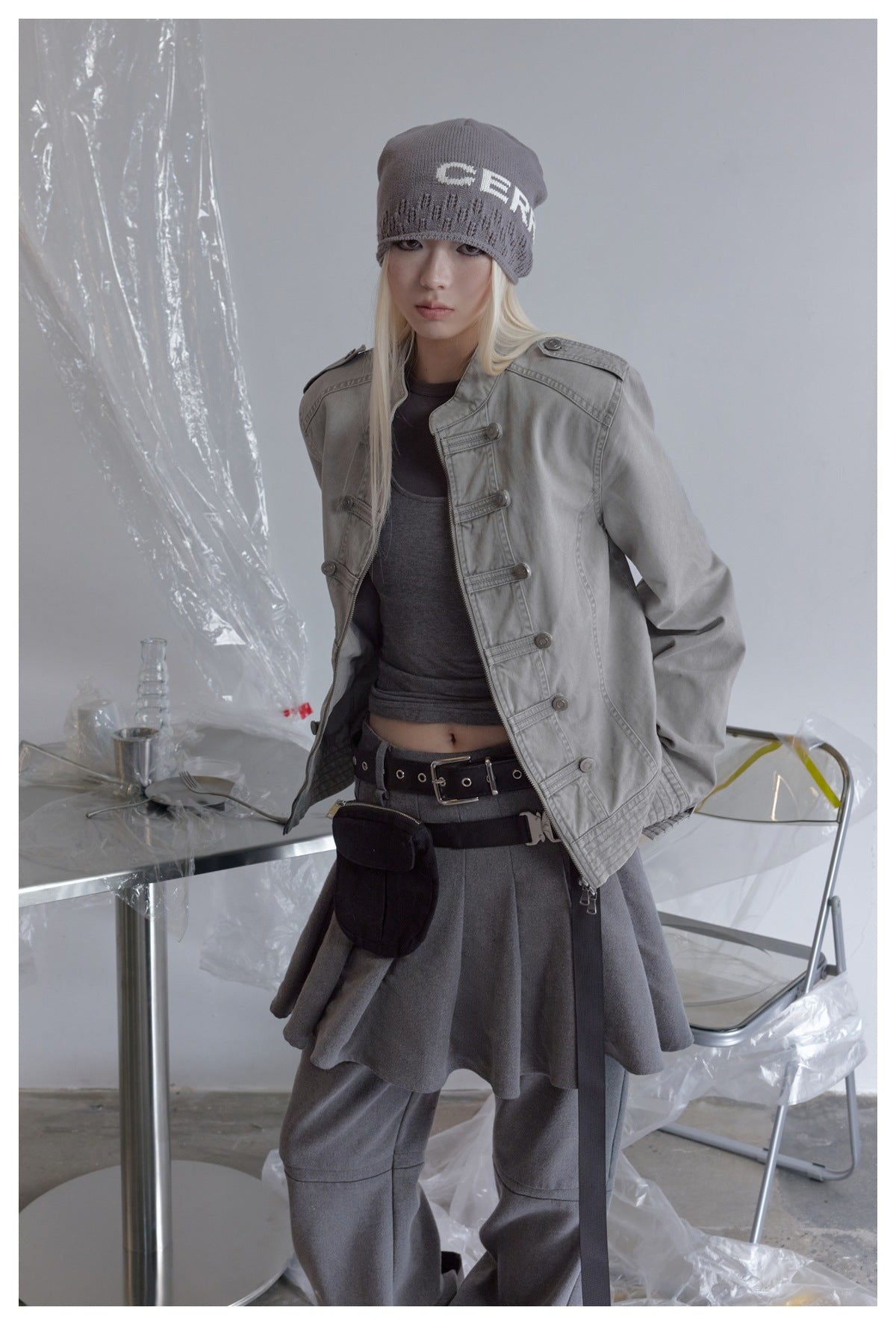 Model wearing open gray military-style zip jacket with braided front bars, over a gray tank top, with a beanie and belt bag