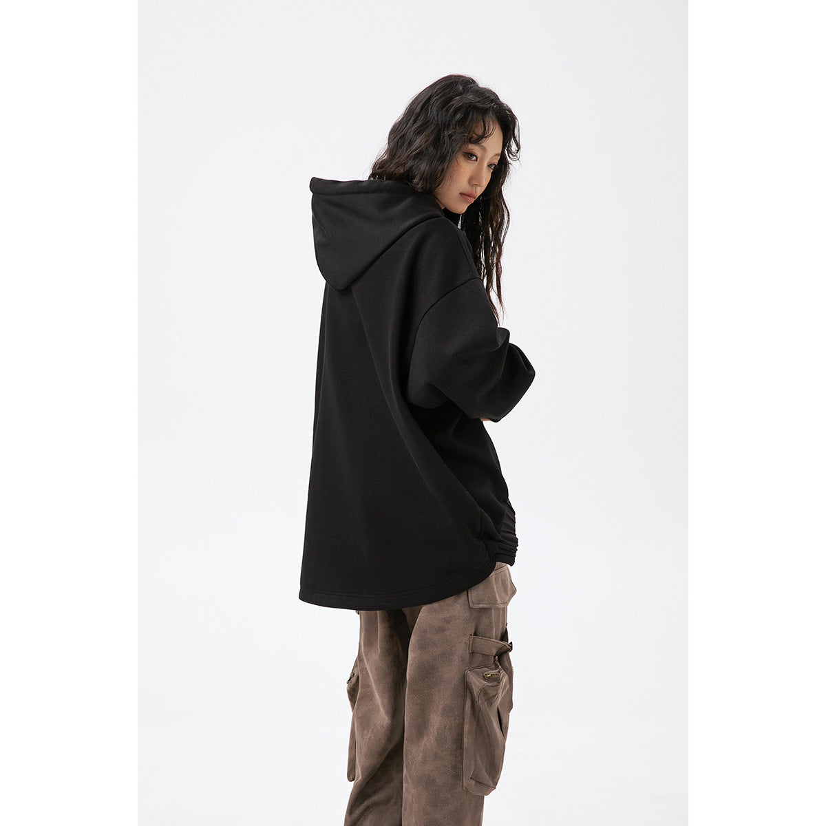 Model in profile wearing an oversized black hoodie, showing its plain hood and back, with brown cargo pants.