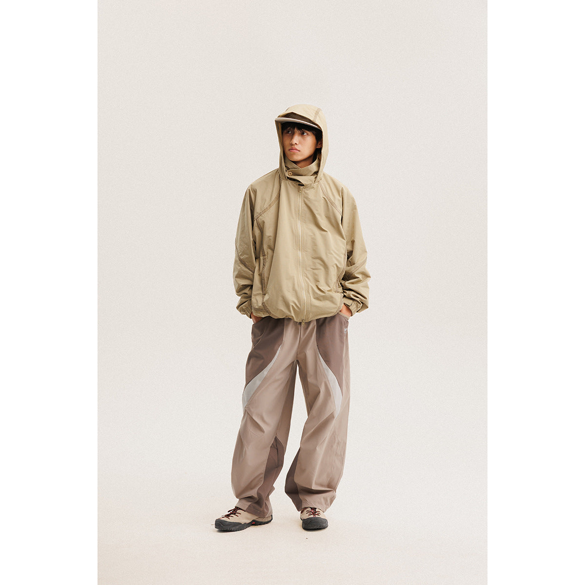 Model wearing a beige hooded zip jacket and loose taupe pants against a plain light background.