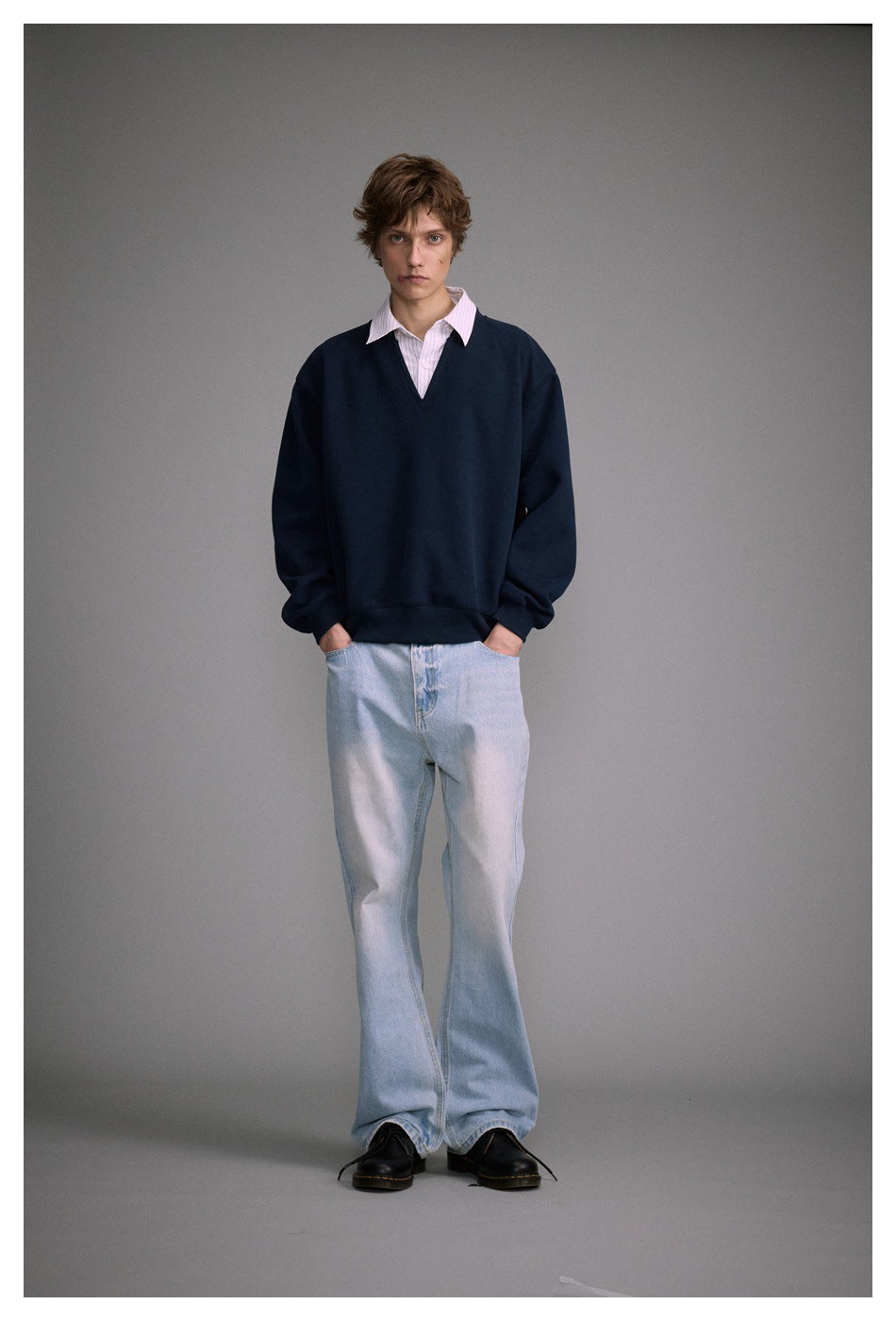 Full-length front view of a model wearing a navy V-neck sweatshirt over a pale pink striped shirt and light-wash jeans.