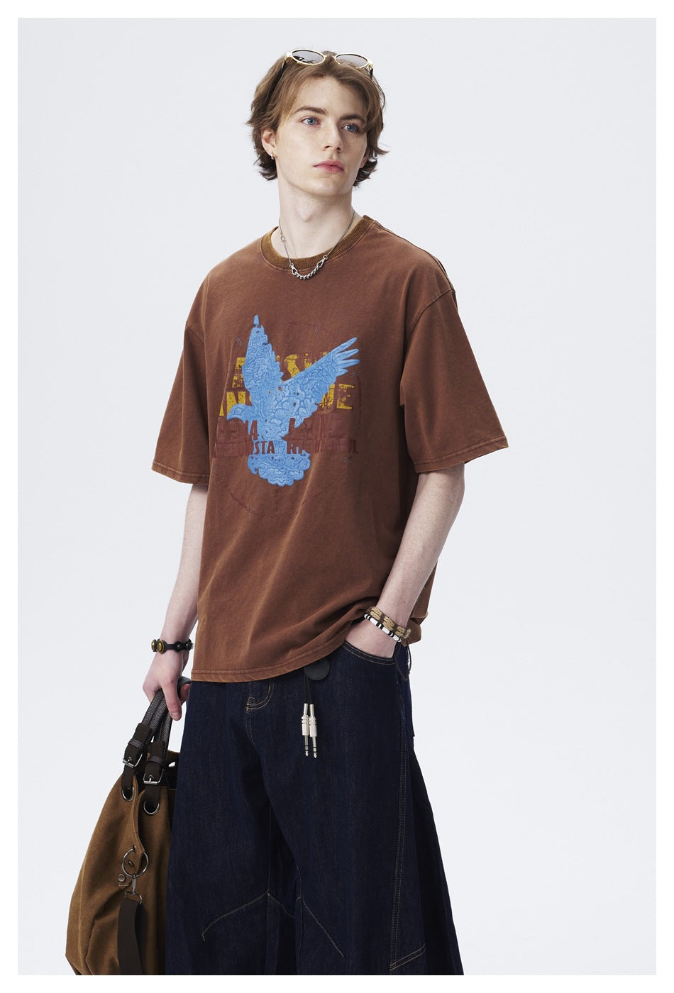 Model wearing a brown oversized T-shirt with a blue eagle graphic, shown at a slight angle with dark jeans.
