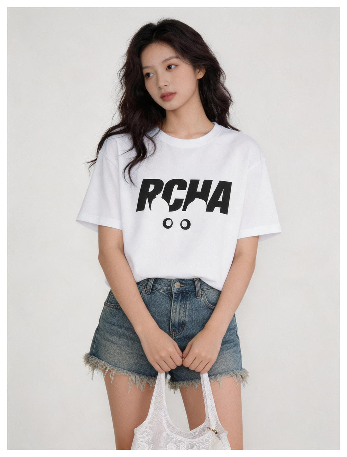 Woman wearing a white relaxed-fit RCHA graphic T-shirt with frayed denim shorts, holding a white mesh bag