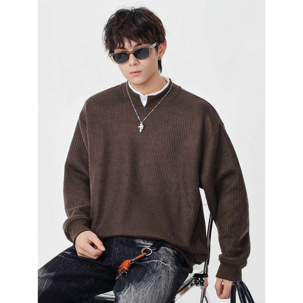 Seated model wearing a dark brown ribbed sweatshirt, sunglasses and a pendant necklace.