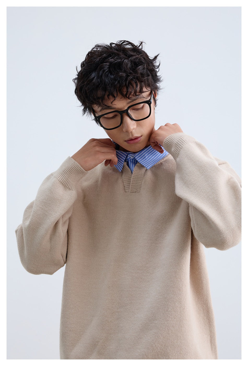 Model wearing a loose beige V-notch sweater over a blue-and-white striped shirt, adjusting the collar.